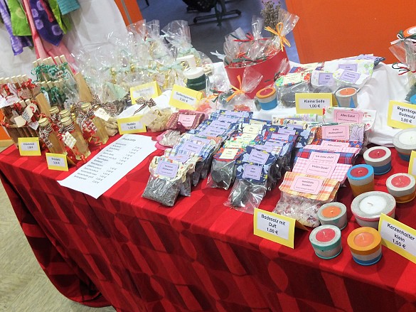 Das Bild zeigt einen Verkaufsstand mit vielen unterschiedlichen Produkten wie Seifen, Ölen, Handtüchern und Kerzenhaltern auf einer roten Tischdecke.
