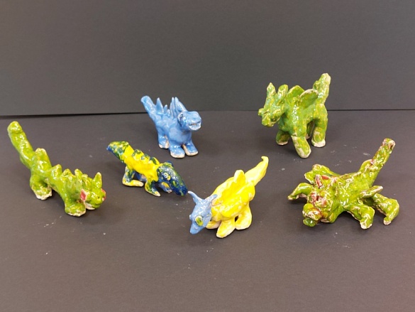 Sechs kleine Drachen aus Ton hinter einem schwarzen Hintergrund. Die Tondrachen sind in unterschiedlichen Farben angemalt (blau, gelb, grün und rot). 