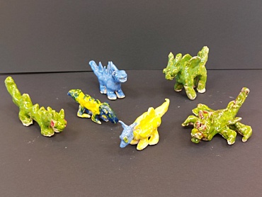 Sechs kleine Drachen aus Ton hinter einem schwarzen Hintergrund. Die Tondrachen sind in unterschiedlichen Farben angemalt (blau, gelb, grün und rot). 