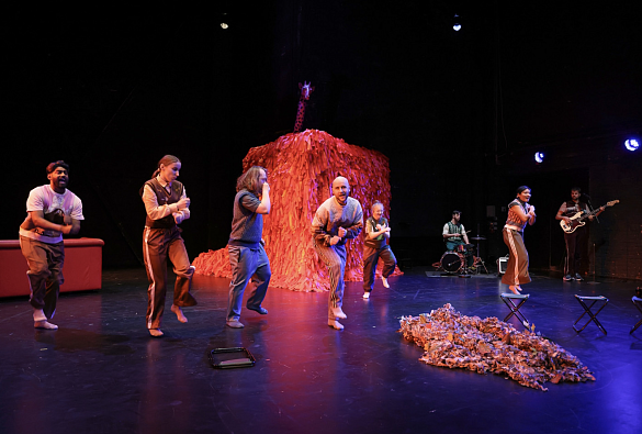 Acht Schauspieler:innen stehen auf der Bühne und tanzen. Ein Schauspieler sitzt am Schlagzeug. Ein Schauspieler spielt Gitarre. Im Hintergrund ein roter Kasten, der ein Haus darstellen soll. 