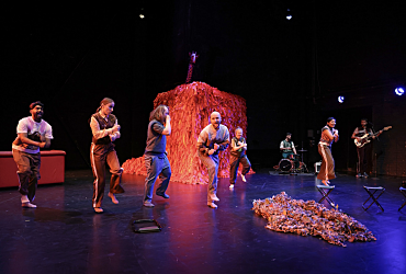 Acht Schauspieler:innen stehen auf der Bühne und tanzen. Ein Schauspieler sitzt am Schlagzeug. Ein Schauspieler spielt Gitarre. Im Hintergrund ein roter Kasten, der ein Haus darstellen soll. 