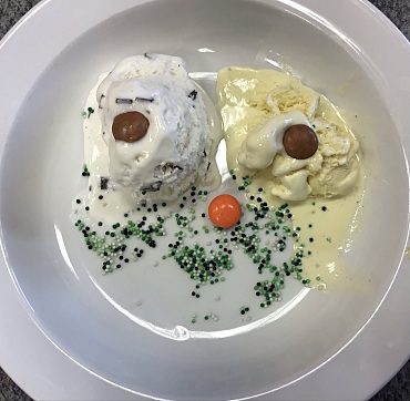 Zwei Eiskugeln auf einem Teller, auf denen jeweils ein brauner Schokodrops liegt. Mittig auf dem Tellerboden ein orangefarbiger Schokodrops. Darunter viele kleine Zuckerkügelchen in Grüntönen. Es ergibt sich der Eindruck eines Gesichtes mit Augen, Nase und Mund.  