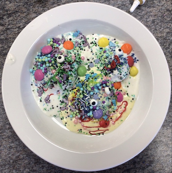 Ein Teller mit zwei Eiskugel, die unter bunten Schokolinsen, Augen aus Zuckerguss, bunten Streuseln in unterschiedlichen Größen, kleinen Zuckersternen sowie Lebensmittelfarbestreifen verdeckt sind. 