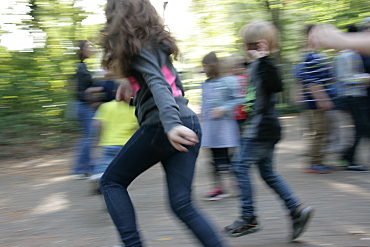 Auf dem Bild sieht man Kinder, die im Kreis über den Schulhof laufen, im Vordergrund ein Mädchen. Das Bild wurde in Bewegung aufgenommen, sodass der Hintergrund verwackelt ist.