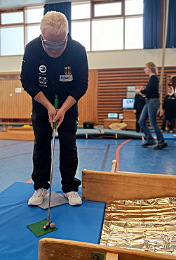 Ein Mann trägt eine Augenbinde. Er hält einen Minigolfschläger in der Hand und legt diesen an den Minigolfball an. Vor ihm ist eine Minigolfbahn aufgebaut. 