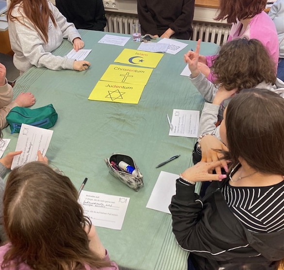 Schüler:innen sitzen gemeinsam am Gruppentisch. Auf dem Gruppentisch sind gelbe Schilder zu sehen mit der Aufschrift: Christentum, Judentum und Islam. 