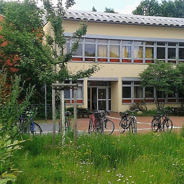 Das Bild zeigt im Hintergrund das hellgelbe Schulgebäude der Georg-Droste-Schule. Im Vordergrund sieht man mehrere Fahrräder an Fahrradständen und eine bunte Blumenwiese.