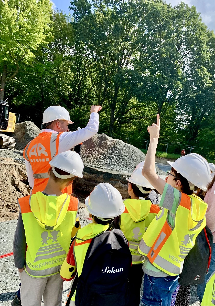 Vier Schüler:innen mit gelber Warnweste und Bauhelm lassen sich vom Bauleiter die Baustelle erklären. 