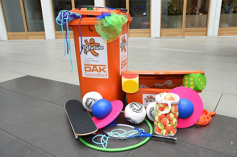 Auf dem Bild sieht man eine orangefarbene Spieletonne mit dem Loge der fit4future-Tonne, aus der Springseile und bunte T&uuml;cher herausragen. Vor der Tonne liegen 3 Fu&szlig;b&auml;lle, ein Tennisschl&auml;ger, ein Skateboard, zwei H&uuml;pfb&auml;lle, viele Tennisb&auml;lle in einem Plastikzylinder, ein Hoolahoopreifen und ein Wurfspiel auf dem Boden.  