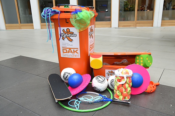 Auf dem Bild sieht man eine orangefarbene Spieletonne mit dem Loge der fit4future-Tonne, aus der Springseile und bunte Tücher herausragen. Vor der Tonne liegen 3 Fußbälle, ein Tennisschläger, ein Skateboard, zwei Hüpfbälle, viele Tennisbälle in einem Plastikzylinder, ein Hoolahoopreifen und ein Wurfspiel auf dem Boden.  