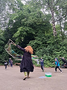 Eine Lehrerin macht Riesenseifenblasen. Drei Kinder rennen an den Seifenblasen vorbei. 
