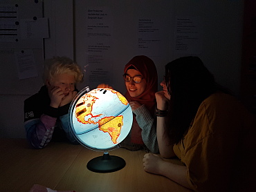 Auf diesem Bild sind drei Schüler*innen unserer Schule zu sehen. Sie sitzen gemeinsam an einem Gruppentisch und schauen auf einen Globus. 