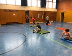 Drei Schüler:innen beim Goalballspielen. Eine Schülerin wirft den Ball, die anderen sitzen auf der Matte. 