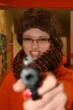 Zu sehen ist eine junge Frau mit Brille, die eine dunkelbraune Kurzhaarperrückte trägt und die Zähne fletscht. Sie schaut direkt in die Kamera und richtet eine Spielzeugpistole mit ausgestrecktem Arm nach vorne. Ihr Pulli ist orangefarben, ihr dicker Schal schwarz orange gemustert.