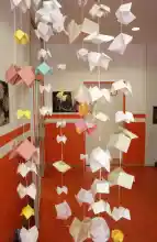 Zu sehen sind lange Girlanden, die an der Decke des Foyers befestigt sind und aus vielen aufgefädelten gefalteten Himmel und Hölle Papieren bestehen.