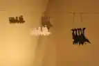 Zu sehen ist ein abgedunkelter Raum, in dem schwarz und weiße ausgeschnittene Gänsefiguren an einer Leine hänge. Diese werden von einer Lampe angestrahlt, sodass man den Schatten der Figuren an der Wand dahinter sehen kann.
