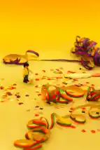 Man sieht Luftschlangen und Konfetti auf hellgelben Untergrund. Auf der linkten Seite befindet sich eine kleine Playmobilfigur, der Hintergrund ist dunkelgelb.