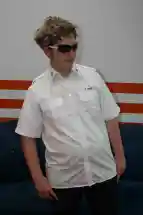 Zu sehen ist ein junger Mann in einem weißen Hemd, der eine dunkle Sonnenbrille trägt und selbstbewusst nach rechts schaut. Die Hände sind in die Hosentaschen gesteckt. Seine Haare sind blond gelockt und etwas länger (Perrücke). Er steht vor einem blauen Sofa, das sich vor einer orange weiß gestreiften Wand befindet.