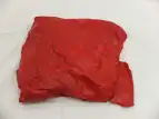 Zu sehen ist ein rot angemaltes Gipsobjekt, was aus einem alten T-Shirt zu einem Quadrat geformt wurde. Die Stoffstrukturen sind teilweise zu erkennen. Es befindet sich auf einem weißen Untergrund aus Stoff.