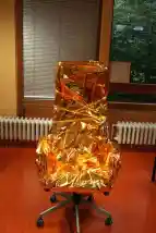 Zu sehen ist ein Schreibtischstuhl mit hoher Lehne, der in einer goldenen Wärmedecke verpackt ist und vor einem Fenster steht.